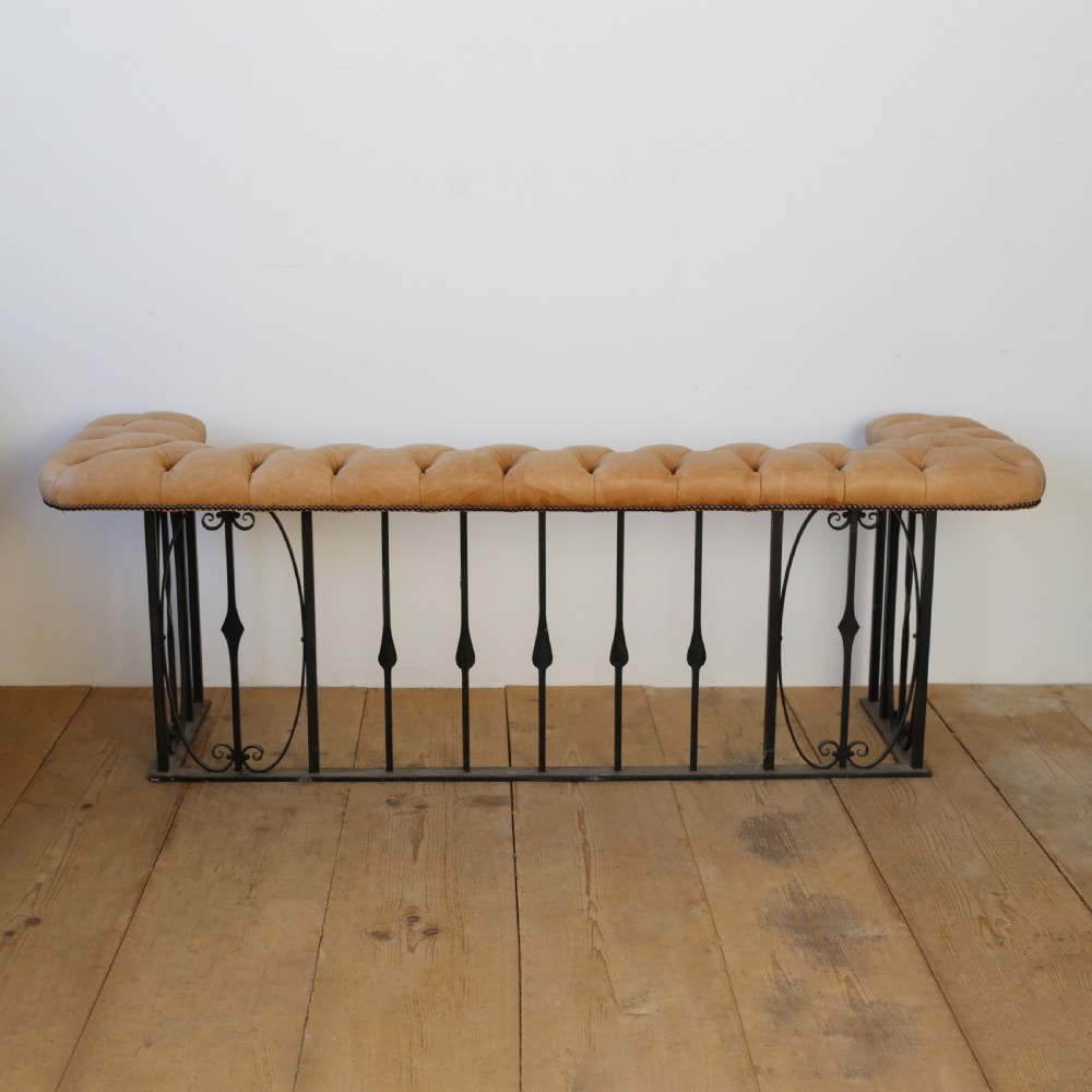 A 1920s English club fender featuring a buttoned tan leather bench top resting on a wrought iron baluster frame with scroll details. View II