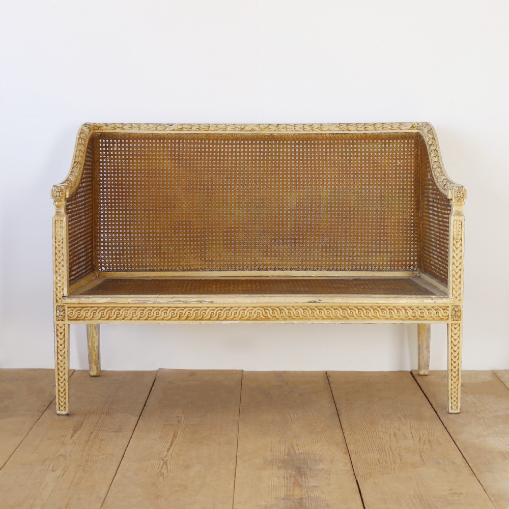 A cream painted wooden settee with a carved guilloche motif frame and woven cane back, side, and seat panels.