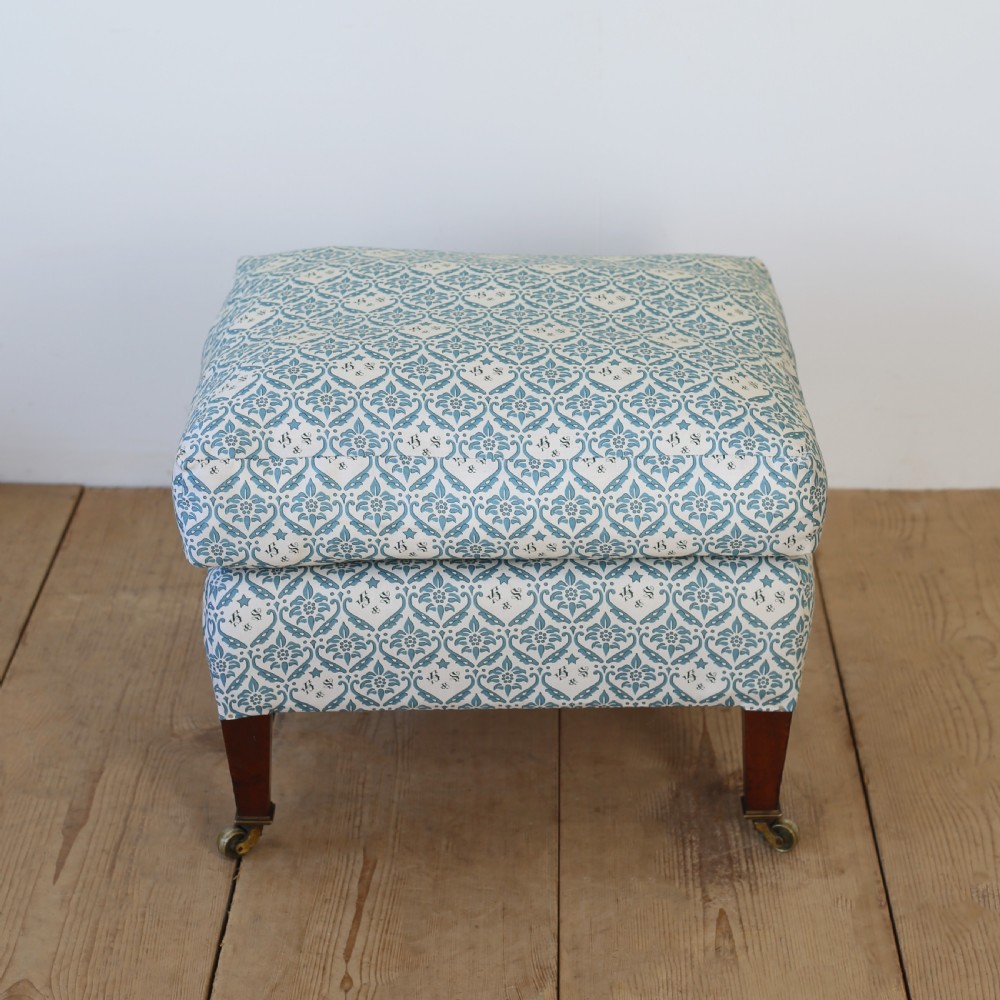 An upholstered rectangular footstool featuring blue and white monogram patterned fabric, tapered wooden legs, and brass castors.