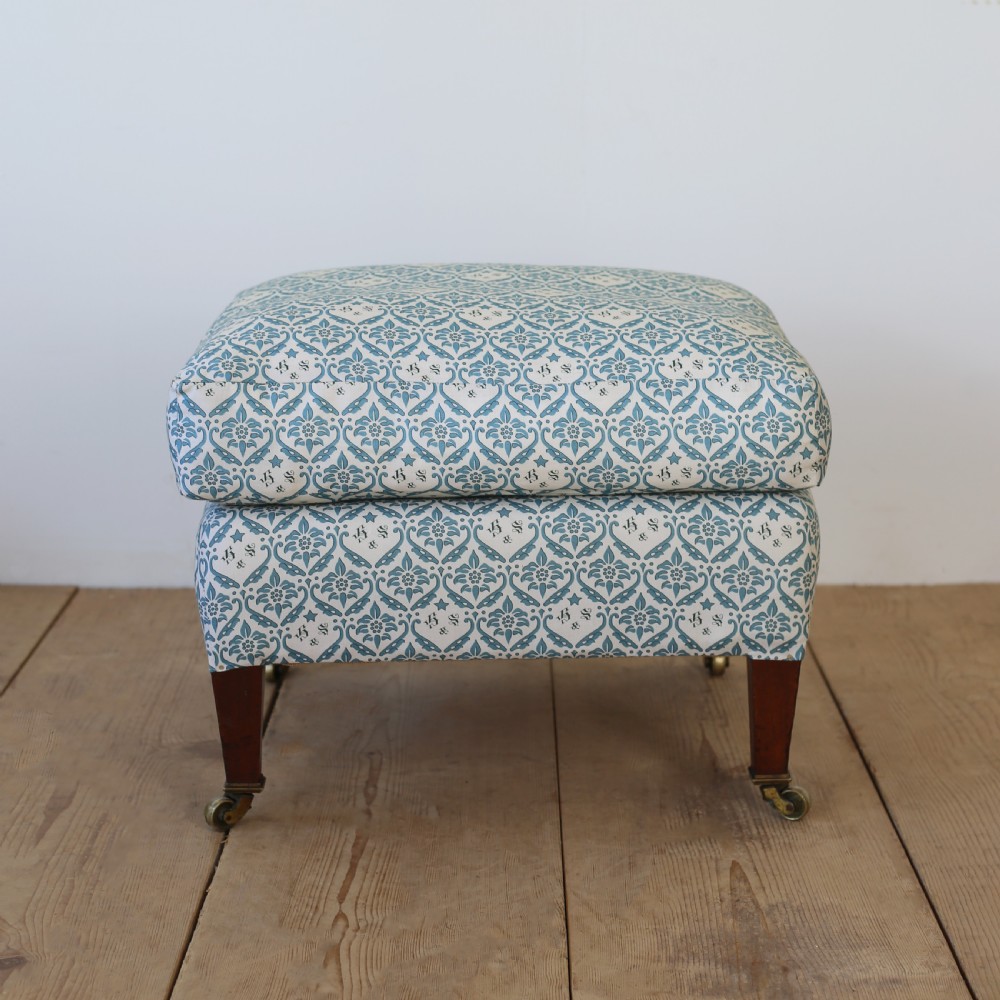 An upholstered rectangular footstool featuring blue and white monogram patterned fabric, tapered wooden legs, and brass castors. View II