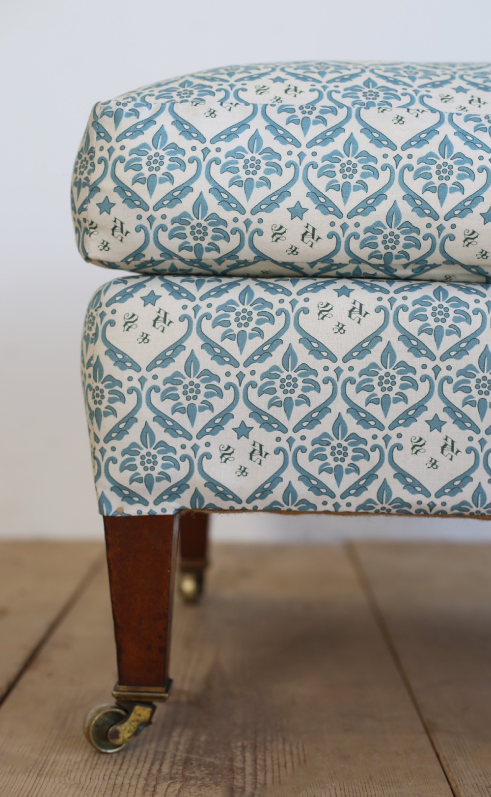 An upholstered rectangular footstool featuring blue and white monogram patterned fabric, tapered wooden legs, and brass castors. View III