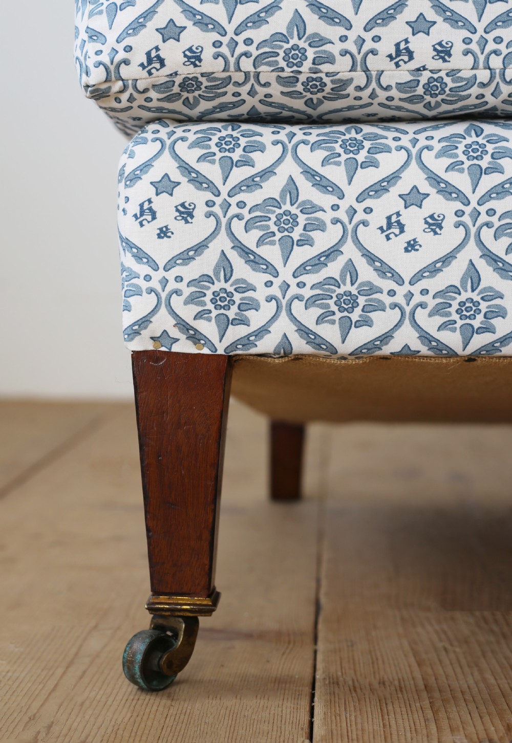 An armless Victorian slipper chair upholstered in blue and white geometric floral patterned fabric with short mahogany legs on brass castors. View II