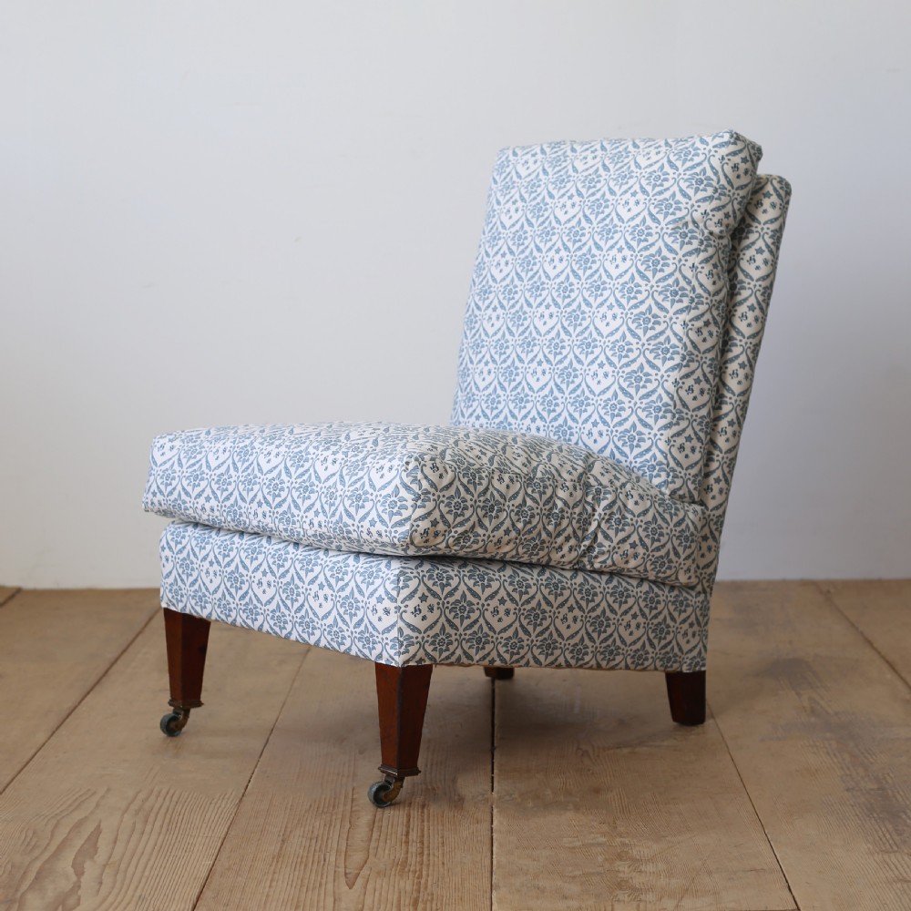 An armless Victorian slipper chair upholstered in blue and white geometric floral patterned fabric with short mahogany legs on brass castors. View V