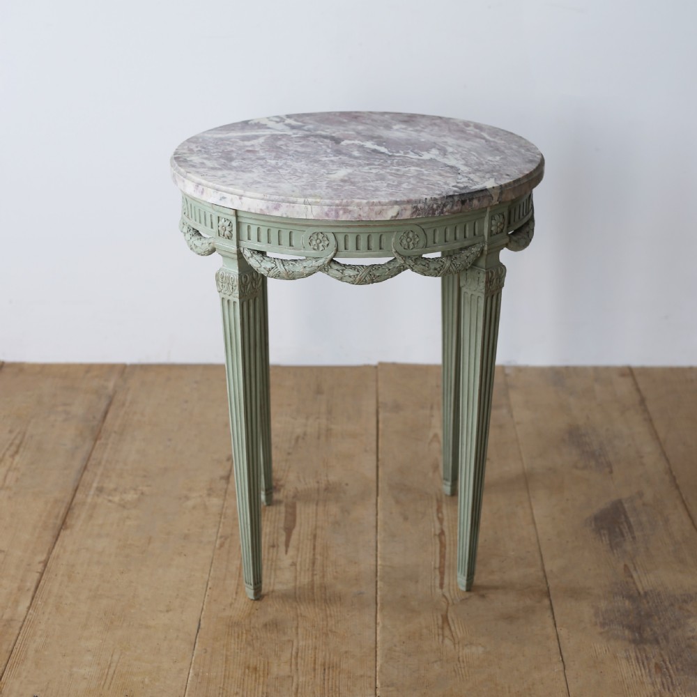 A circular 20th-century gueridon side table with a variegated pink marble top and a sage-green painted wooden frame featuring carved swags and fluted legs.