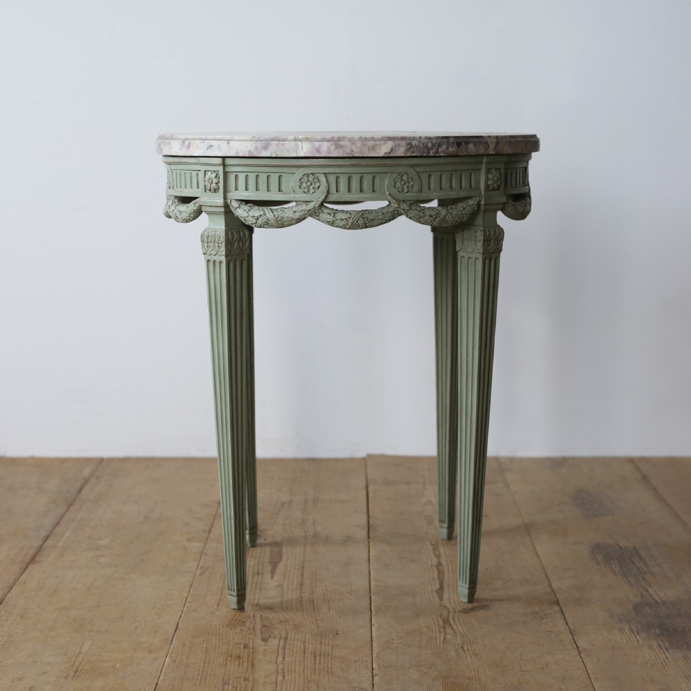A circular 20th-century gueridon side table with a variegated pink marble top and a sage-green painted wooden frame featuring carved swags and fluted legs. View II