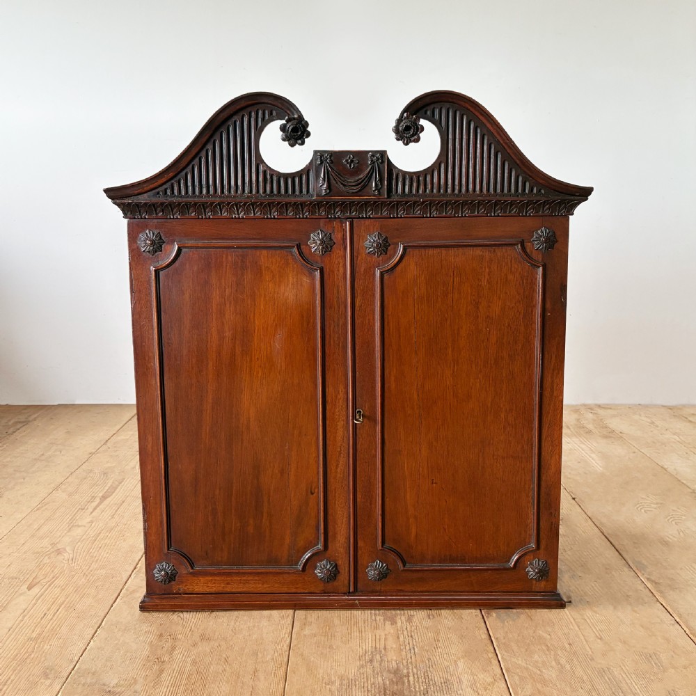 A George II mahogany wall cabinet featuring a carved swan-neck pediment and panelled twin doors with rosette corner accents.