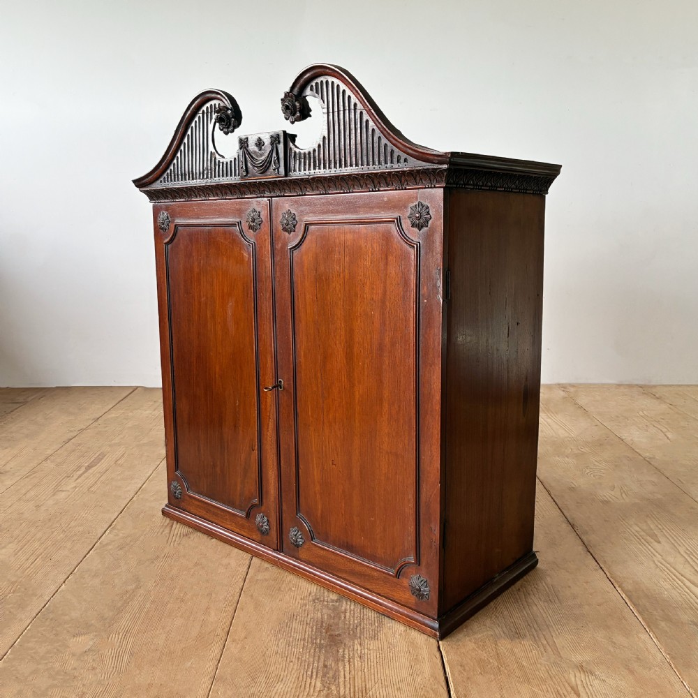 A George II mahogany wall cabinet featuring a carved swan-neck pediment and panelled twin doors with rosette corner accents. View III