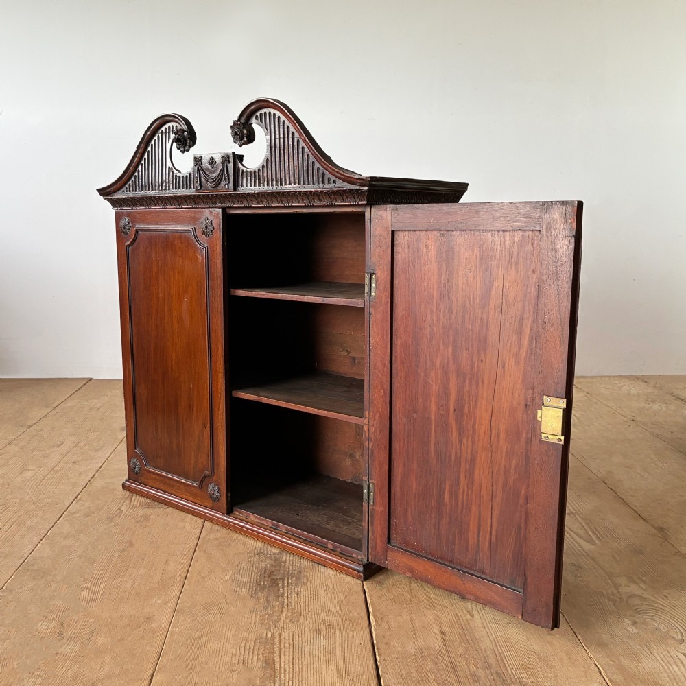 A George II mahogany wall cabinet featuring a carved swan-neck pediment and panelled twin doors with rosette corner accents. View IV