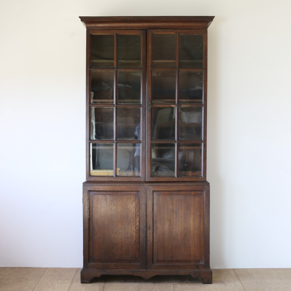 A tall George III dark oak glazed bookcase featuring two upper glass panelled doors over a double cupboard base supported by bracket feet.