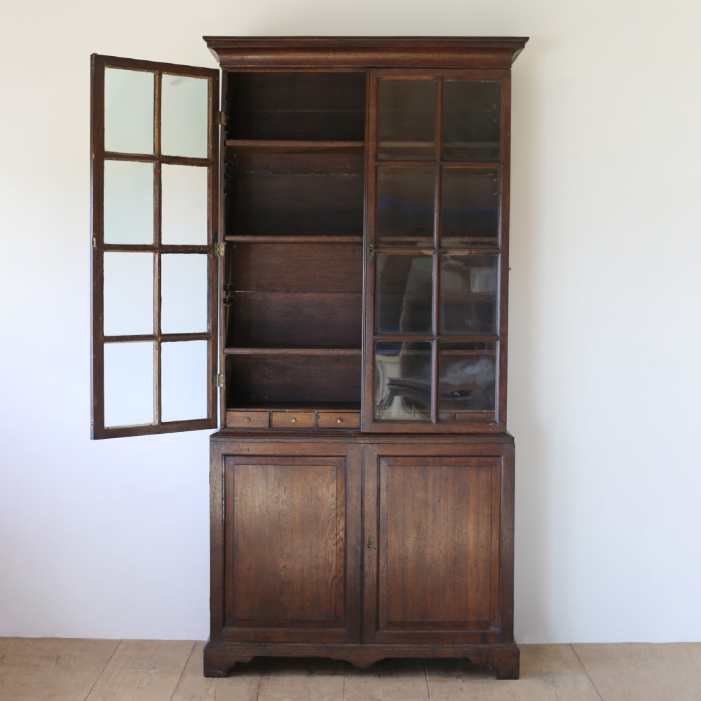 A tall George III dark oak glazed bookcase featuring two upper glass panelled doors over a double cupboard base supported by bracket feet. View II