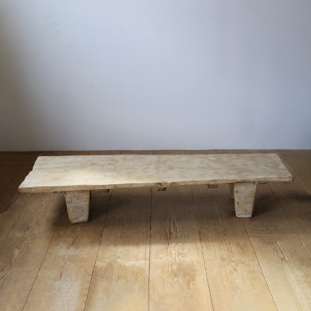 A low-slung, pale wooden Indian daybed featuring a flat rectangular timber surface resting on four heavy, tapered block legs.