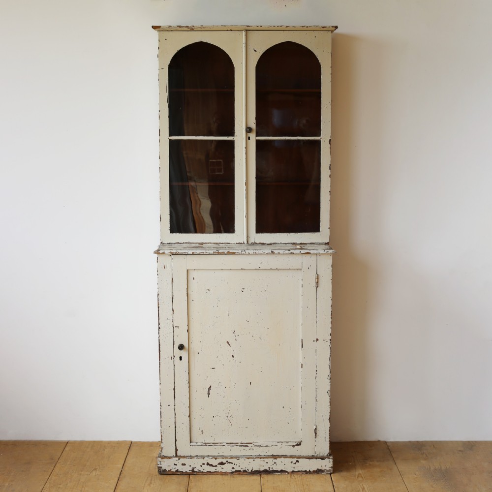 A tall 19th-century cream painted wooden cabinet featuring upper glazed doors with arched glass panels above a solid cupboard base.