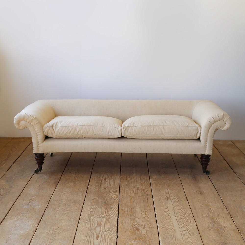 A 19th-century English country house sofa upholstered in cream linen fabric, featuring scrolled arms, two loose seat cushions, and turned mahogany legs with brass castors.