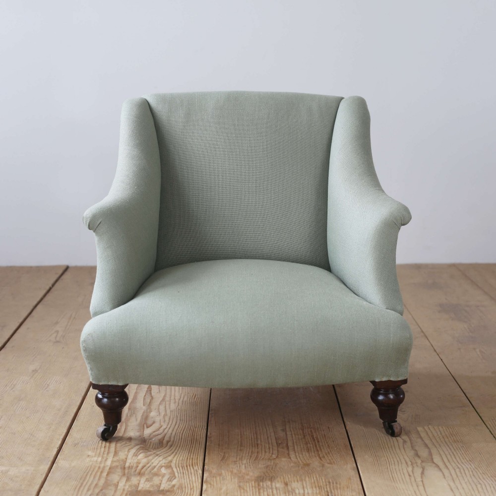 A 19th-century Victorian armchair upholstered in sage green fabric, featuring sloping arms and dark turned wooden front legs with castors.
