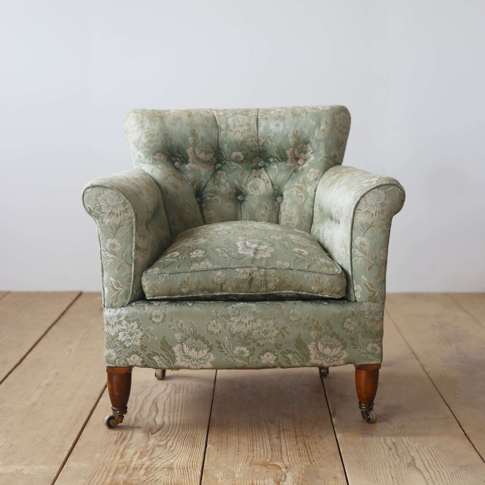 A 19th-century upholstered armchair featuring sage green floral damask fabric, deep button-tufting on the backrest, turned wood front legs, and brass castors.