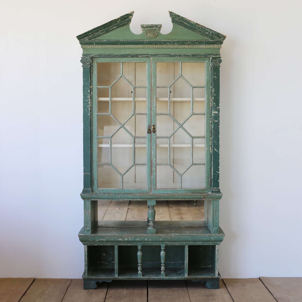 A 19th-century distressed green painted glazed bookcase featuring a broken pediment, astragal glazing doors, and open lower cubby compartments.