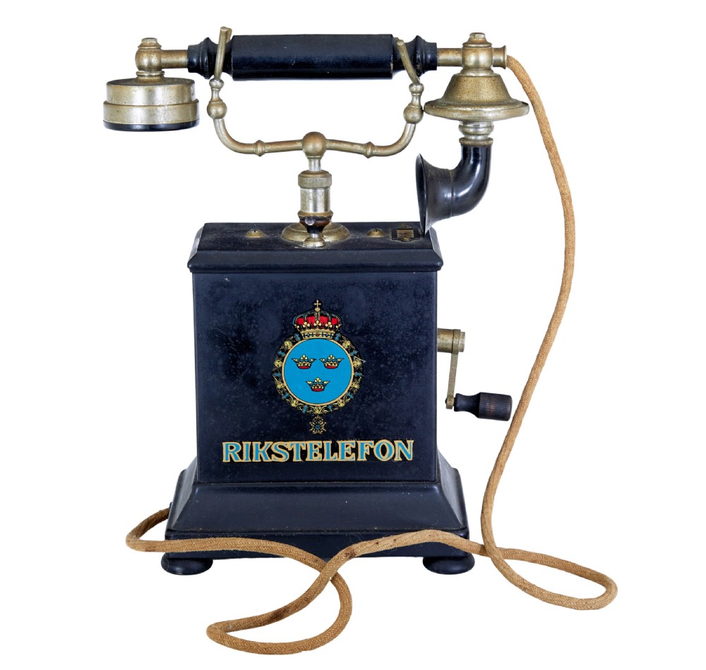 An early twentieth-century black metal Swedish Rikstelefon tabletop telephone with a brass handset cradle, fabric cord, side hand crank, and central blue crown decal. View II