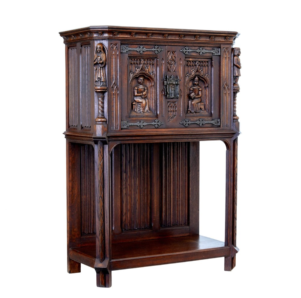 A dark oak Renaissance Revival cupboard featuring carved human figures, gothic arched panels, iron hardware, and an open lower shelf.