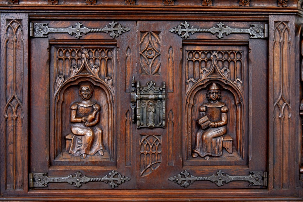 A dark oak Renaissance Revival cupboard featuring carved human figures, gothic arched panels, iron hardware, and an open lower shelf. View VII
