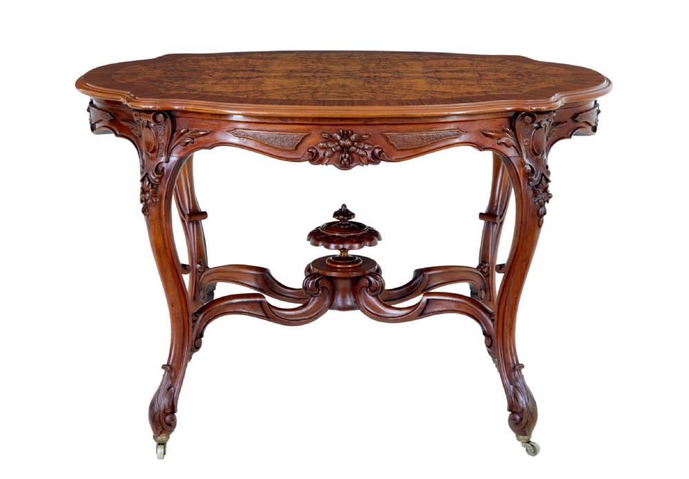 A 19th-century carved walnut occasional table featuring a shaped burr walnut top, ornate floral apron, cabriole legs, and a carved central stretcher finial on castors.