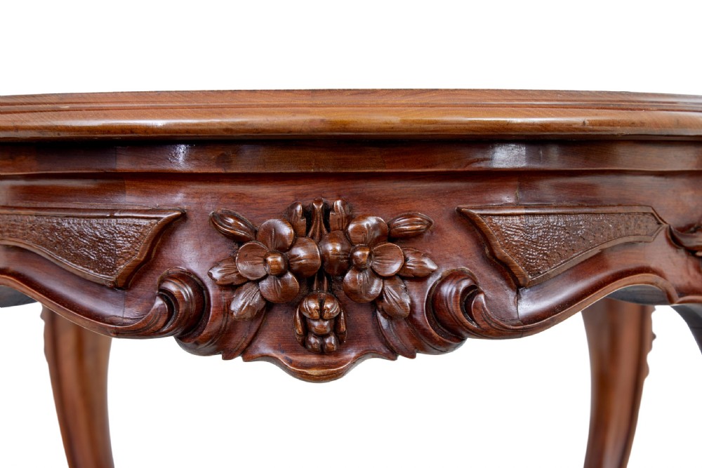 A 19th-century carved walnut occasional table featuring a shaped burr walnut top, ornate floral apron, cabriole legs, and a carved central stretcher finial on castors. View V