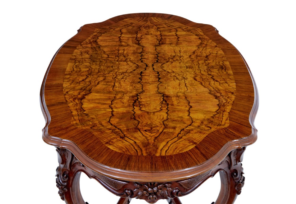 A 19th-century carved walnut occasional table featuring a shaped burr walnut top, ornate floral apron, cabriole legs, and a carved central stretcher finial on castors. View IX