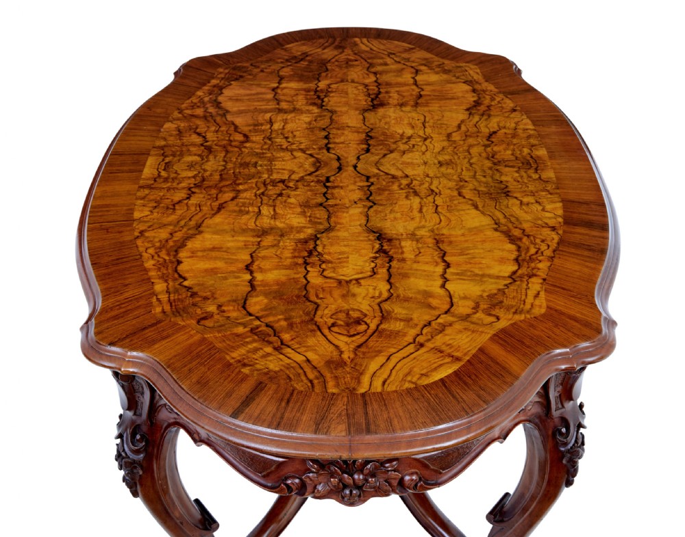 A 19th-century carved walnut occasional table featuring a shaped burr walnut top, ornate floral apron, cabriole legs, and a carved central stretcher finial on castors. View X