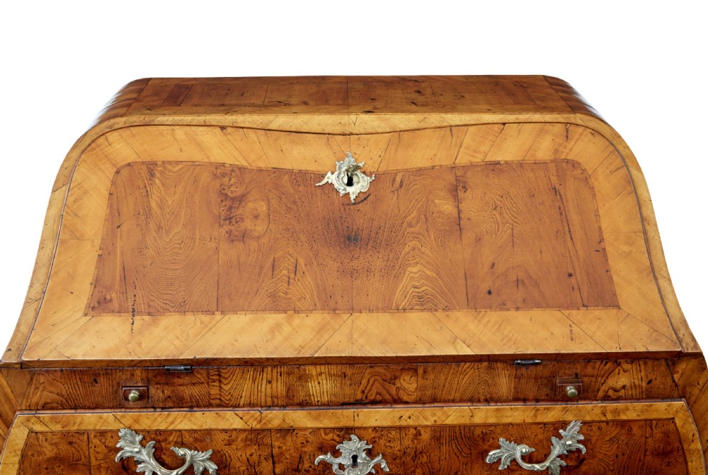 An 18th-century Rococo bureau crafted from yew and elm wood featuring a curved bombe body, slant-front writing slope, three drawers, and decorative cast brass handles. View X