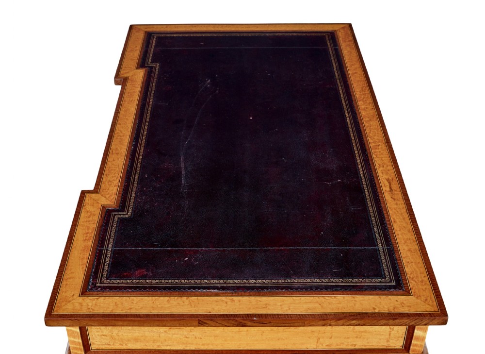 An early 20th-century Sheraton Revival satinwood pedestal desk featuring inlaid mahogany crossbanding, seven drawers with brass handles, and gently splayed legs. View IX