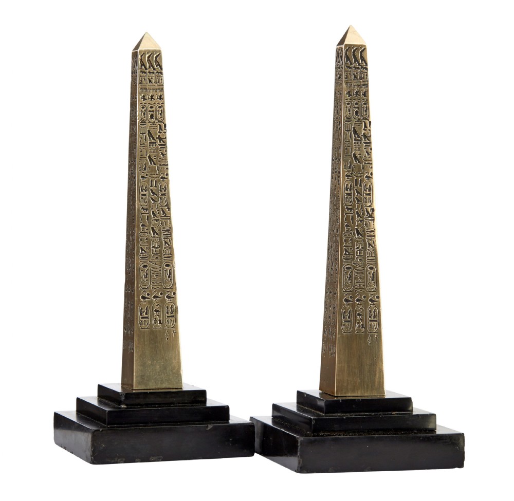 A pair of brass obelisks with incised hieroglyphic carvings mounted on stepped black marble bases.