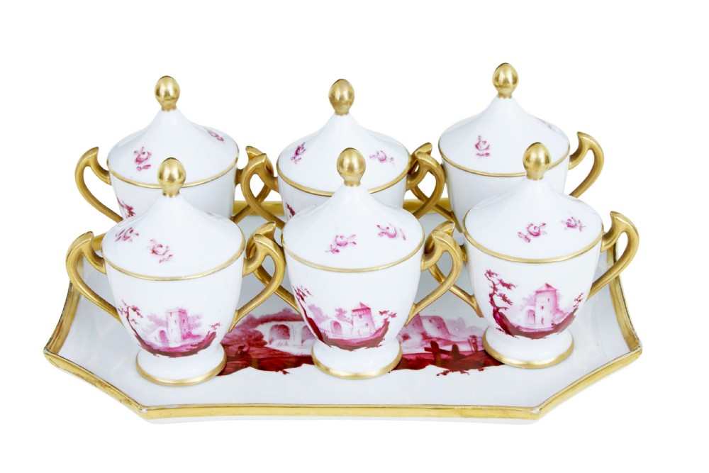 A seven-piece French porcelain dessert set featuring six twin-handled covered cream pots and a matching shaped tray, decorated with pink landscape scenes and gilded handles.