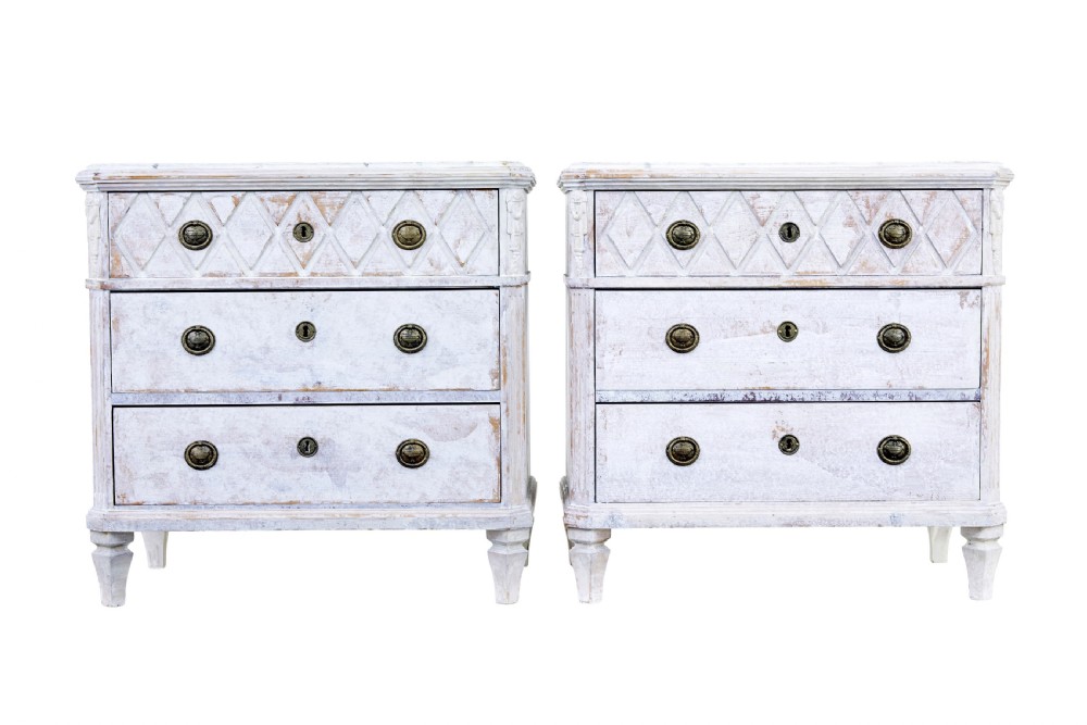 A pair of 19th-century Swedish chests of drawers featuring three drawers each, diamond lattice carvings on the top drawer front, brass handles, and a distressed white paint finish.