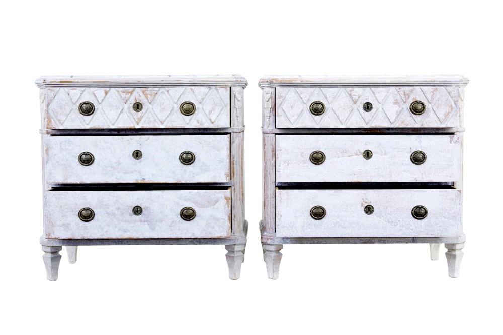 A pair of 19th-century Swedish chests of drawers featuring three drawers each, diamond lattice carvings on the top drawer front, brass handles, and a distressed white paint finish. View II