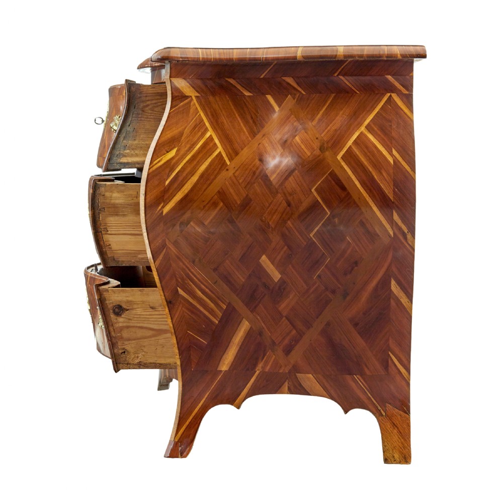An early 18th-century Rococo bombe chest of drawers featuring geometric parquetry inlay in plumwood and three drawers with ornate brass handles. View V