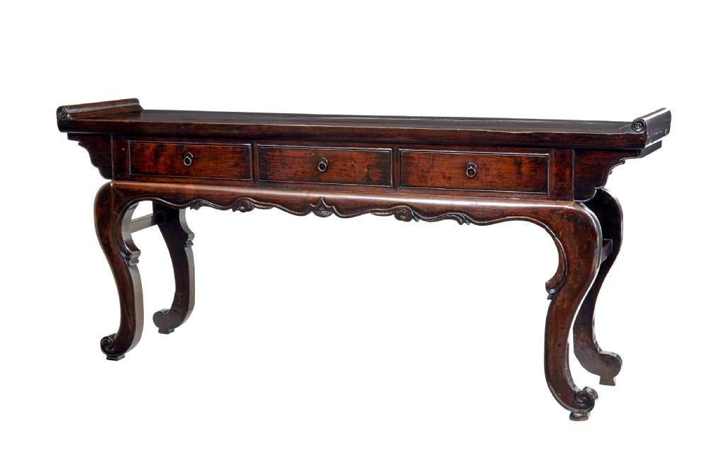 A nineteenth-century dark lacquered Chinese hardwood altar table with three drawers, everted end flanges, carved aprons, and curved legs.