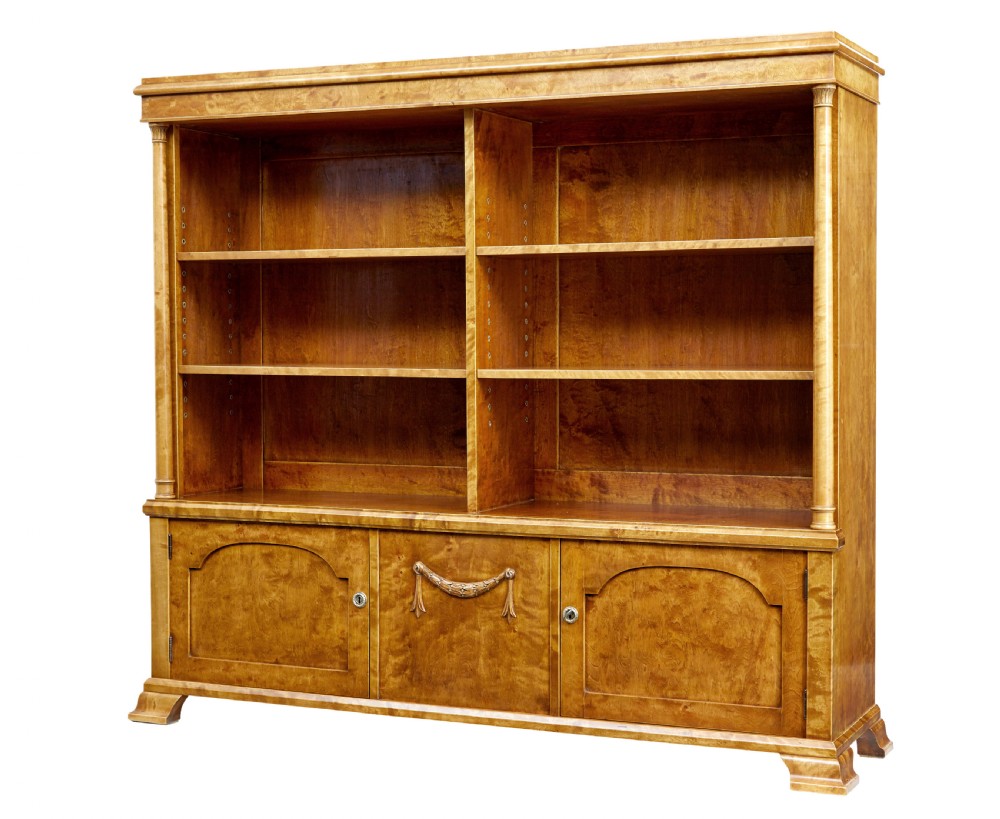 An early twentieth-century Empire Revival satin birch bookcase cabinet featuring open adjustable upper shelving and three lower panelled cupboard doors with a central carved drape motif.