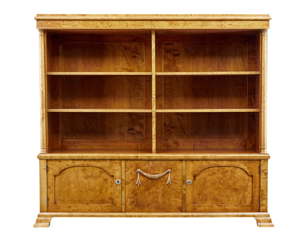 An early twentieth-century Empire Revival satin birch bookcase cabinet featuring open adjustable upper shelving and three lower panelled cupboard doors with a central carved drape motif. View II