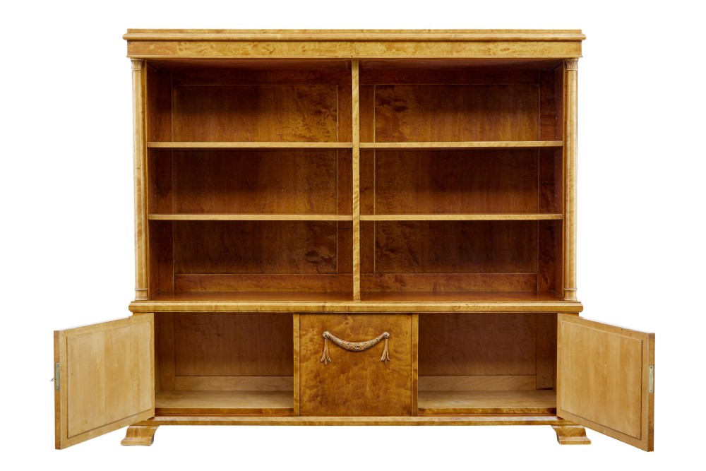 An early twentieth-century Empire Revival satin birch bookcase cabinet featuring open adjustable upper shelving and three lower panelled cupboard doors with a central carved drape motif. View III