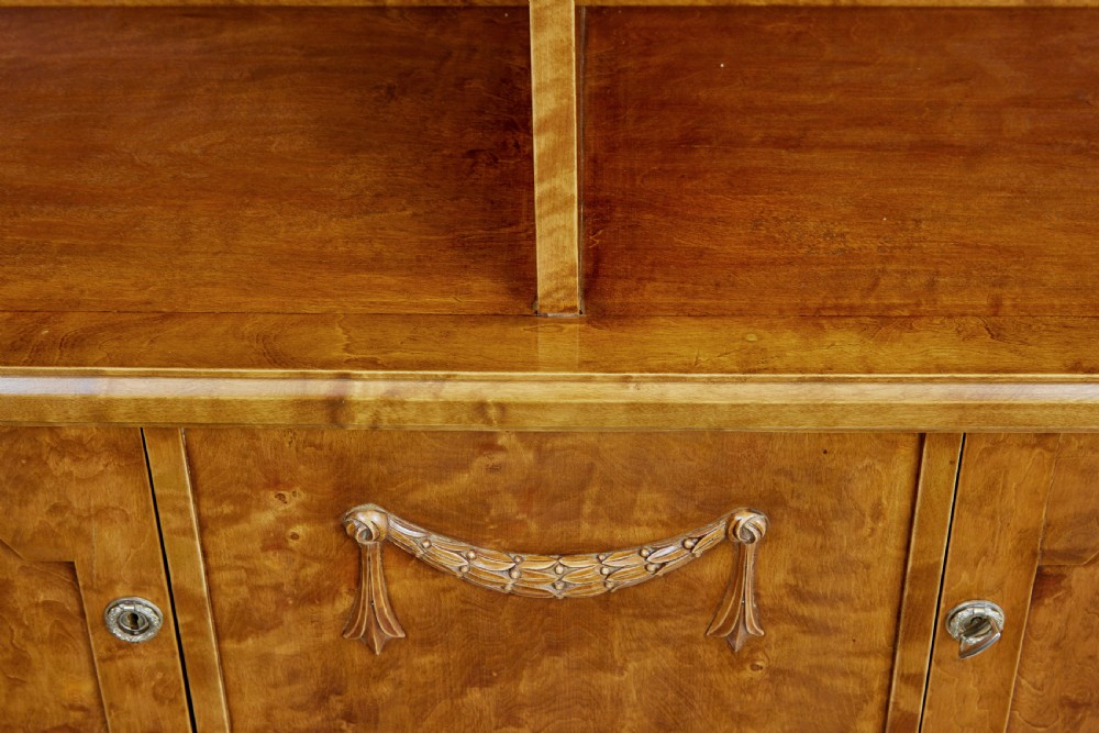 An early twentieth-century Empire Revival satin birch bookcase cabinet featuring open adjustable upper shelving and three lower panelled cupboard doors with a central carved drape motif. View VII
