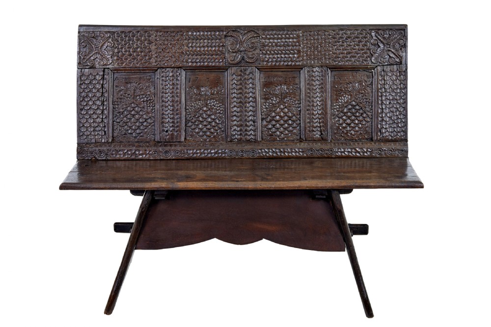 A dark carved oak bench featuring a high panelled back with intricate relief carvings and splayed trestle legs.