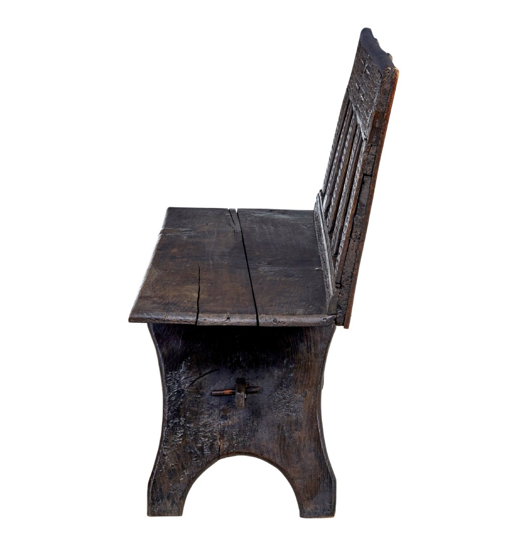 A dark carved oak bench featuring a high panelled back with intricate relief carvings and splayed trestle legs. View II