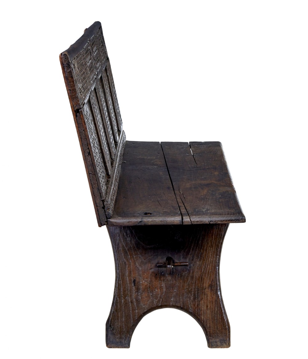A dark carved oak bench featuring a high panelled back with intricate relief carvings and splayed trestle legs. View III
