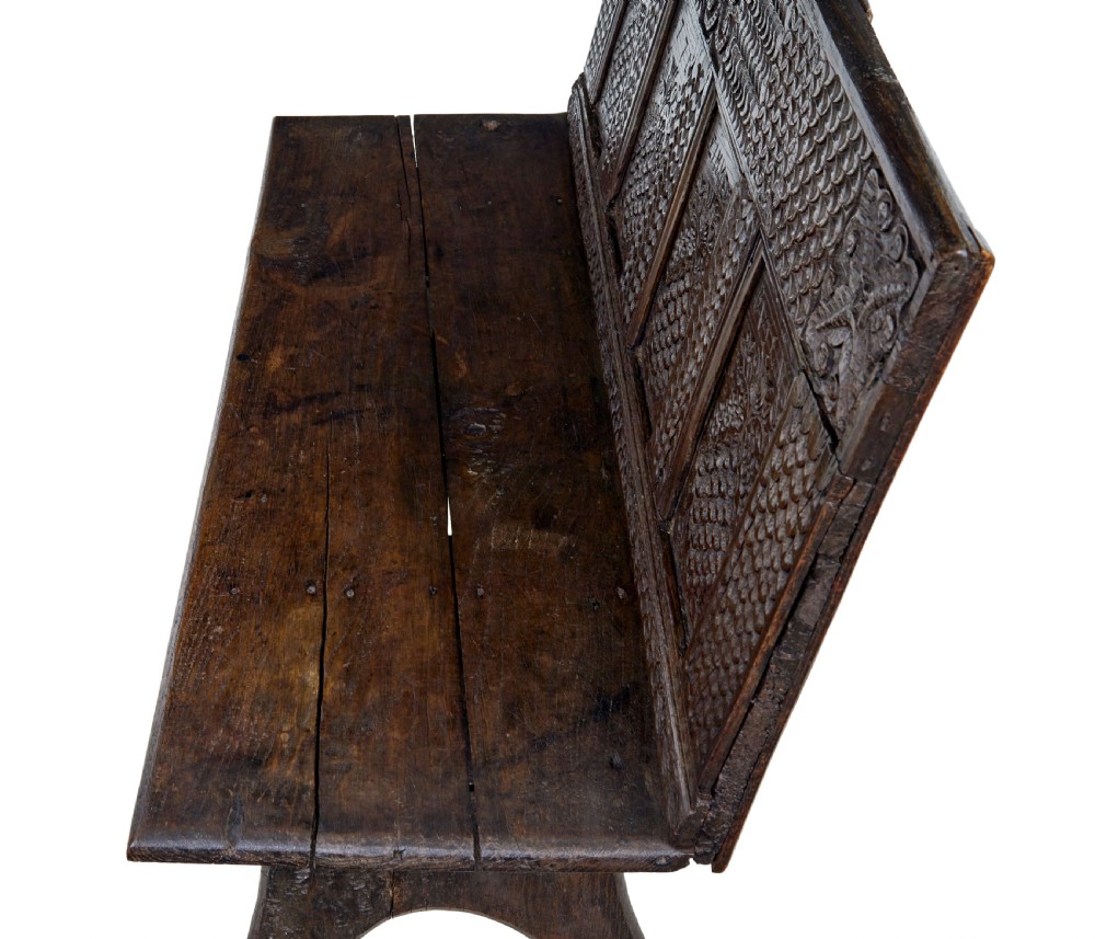 A dark carved oak bench featuring a high panelled back with intricate relief carvings and splayed trestle legs. View V