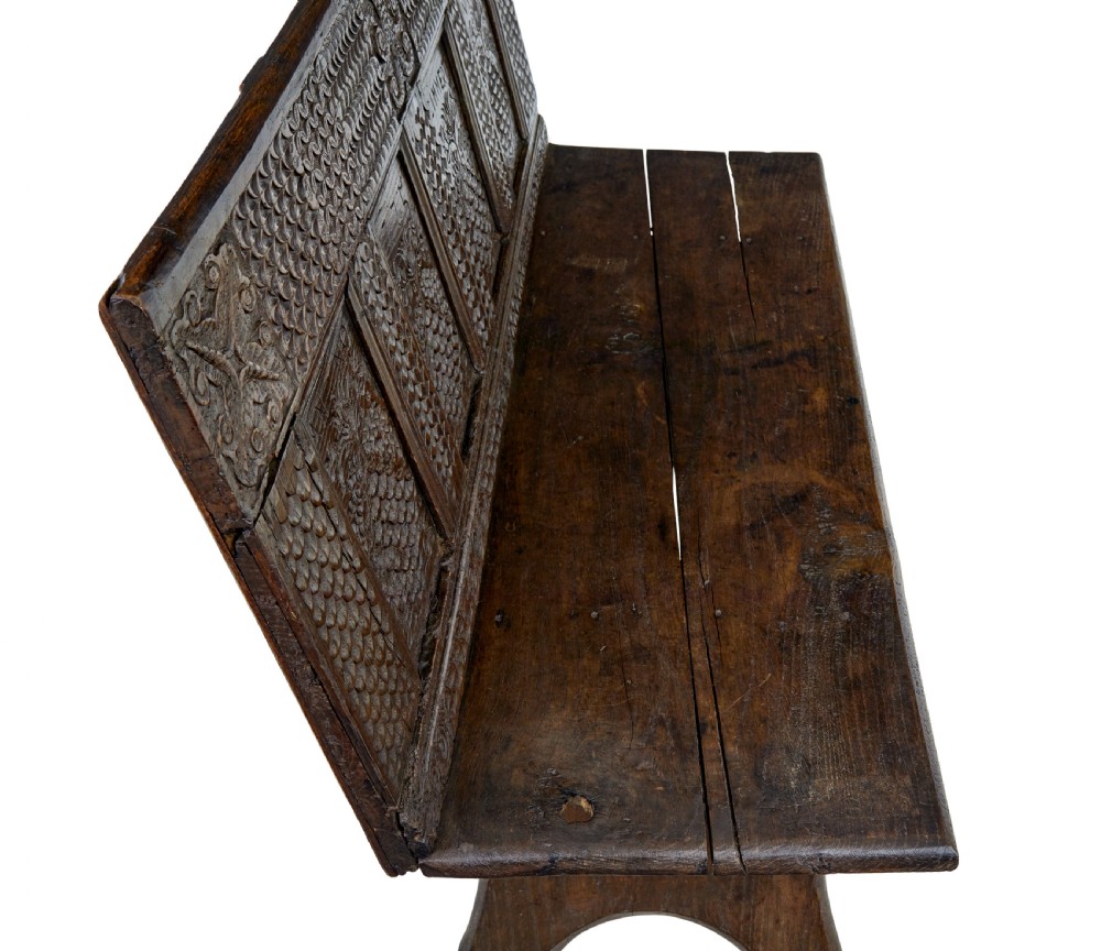 A dark carved oak bench featuring a high panelled back with intricate relief carvings and splayed trestle legs. View VI
