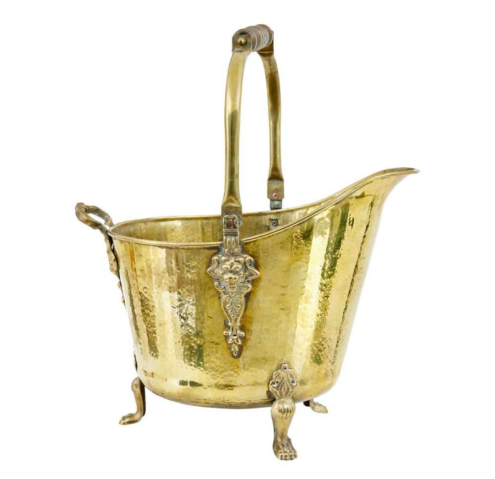 A 19th-century hammered brass helmet coal scuttle with an overhead handle, ornate side mounts, and paw feet.