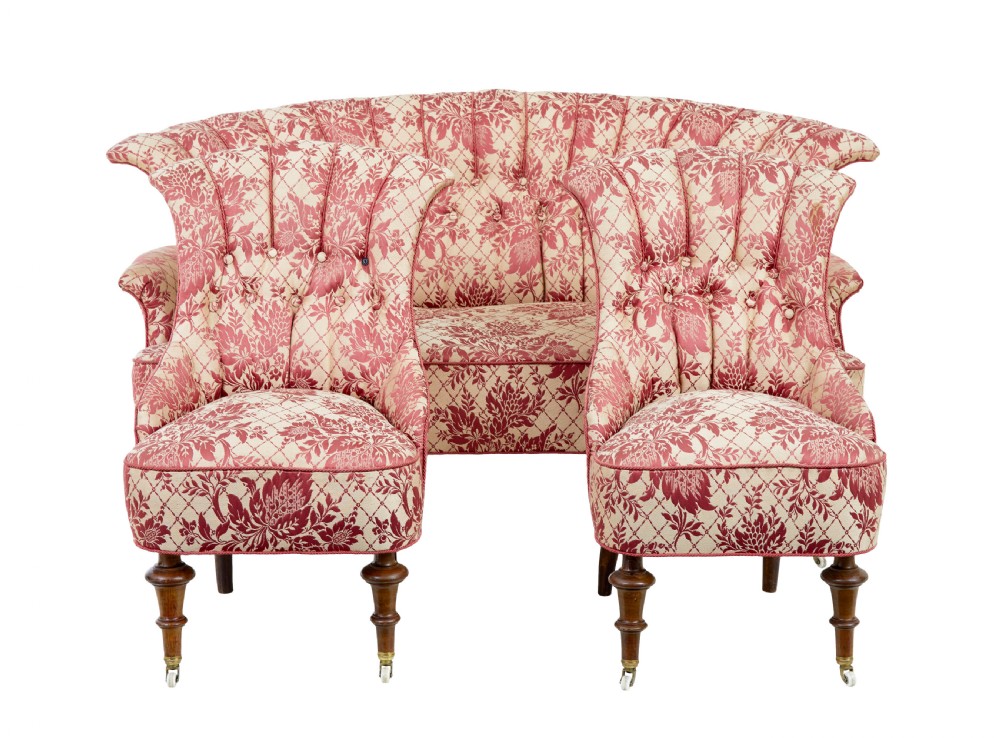 An early 20th century three-piece upholstered salon suite featuring a two-seater settee and two matching armchairs with pink and cream floral damask pattern, button-tufted fluted backs, and turned wooden legs with brass castors.