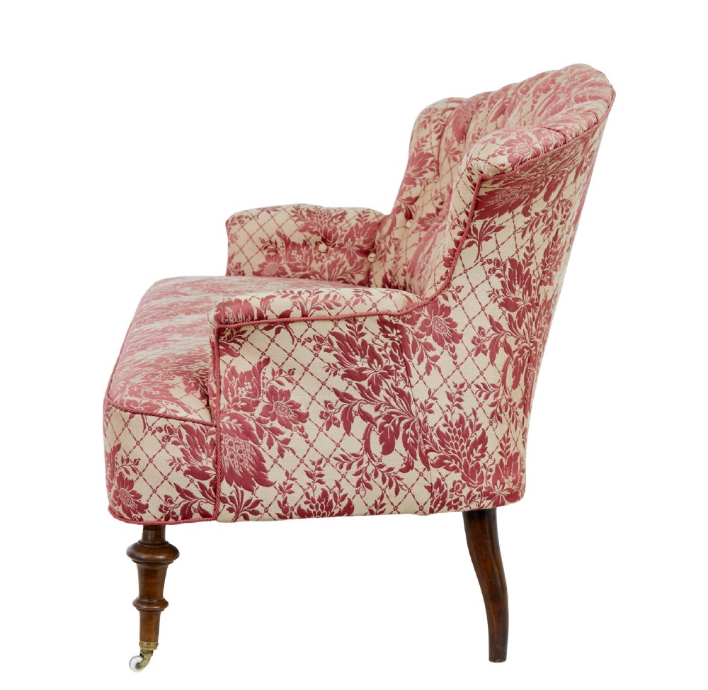 An early 20th century three-piece upholstered salon suite featuring a two-seater settee and two matching armchairs with pink and cream floral damask pattern, button-tufted fluted backs, and turned wooden legs with brass castors. View VI