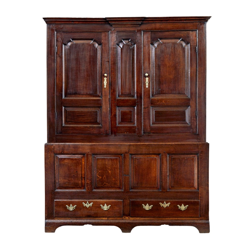 An 18th-century dark oak housekeepers cupboard featuring arched fielded panel doors above a lower section fitted with drawers and brass handles.