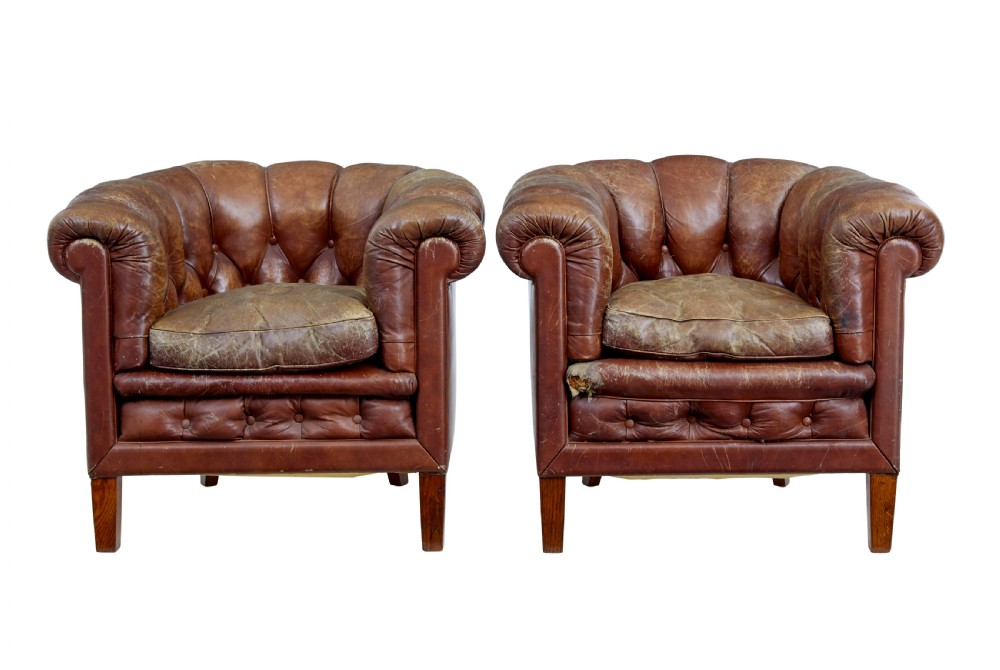 A pair of early 20th-century brown leather club armchairs with deep buttoning, rolled arms, loose seat cushions, and square timber legs.