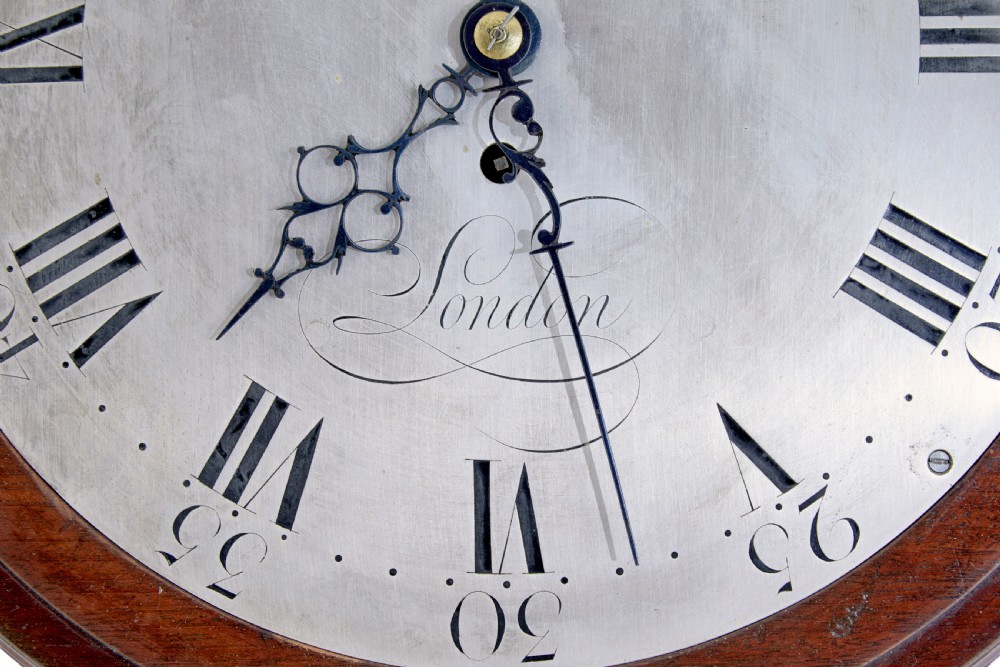 An 18th-century Georgian wall clock featuring an engraved silvered dial with Roman numerals, a brass bezel, and a circular mahogany surround. View V